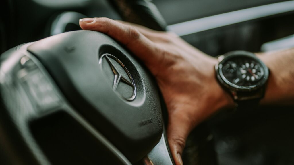 Close-up of a man's hand on a Mercedes steering wheel, wearing a watch.