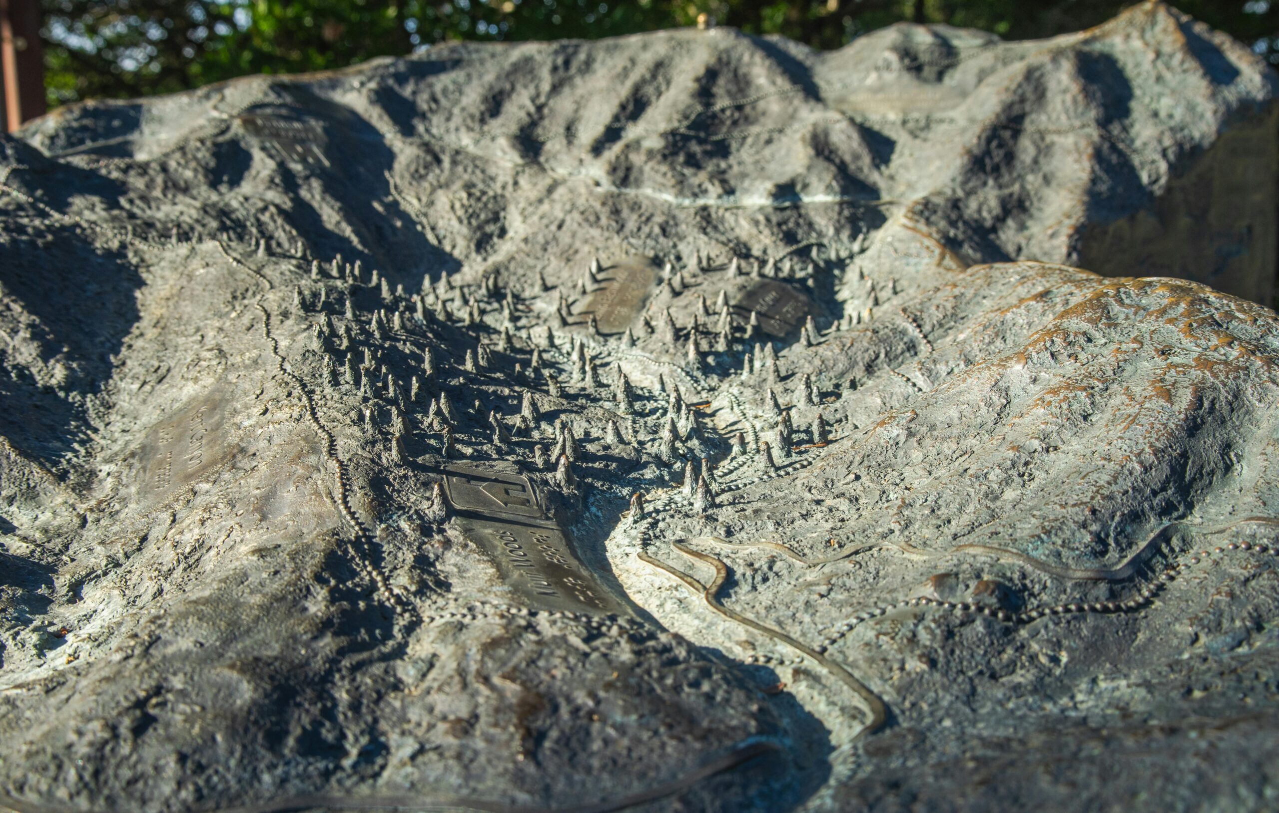 Detailed bronze relief map depicting mountainous terrain with a village and pathways under sunlight.
