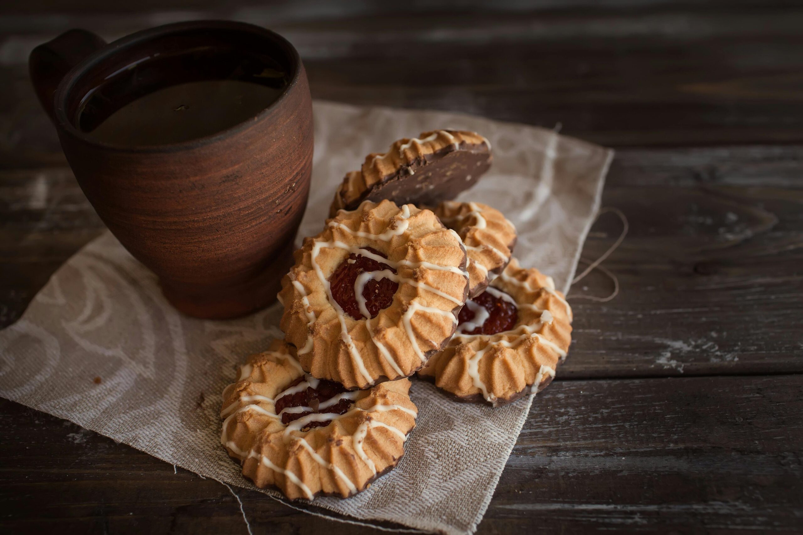 Rustic brown mug with hot beverage and jam-filled cookies on a wooden table, creating a cozy vibe.