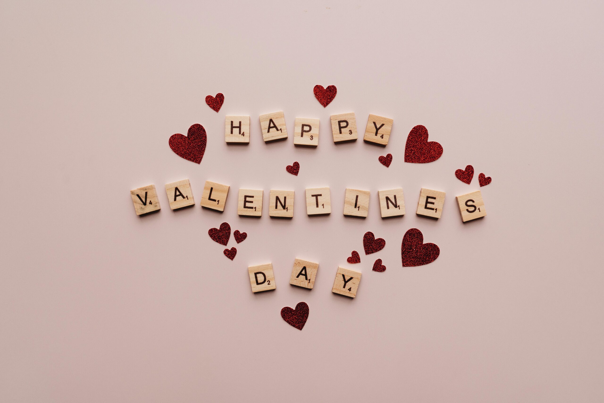 A festive Valentine's Day greeting with Scrabble tiles and red glitter hearts on a soft background.
