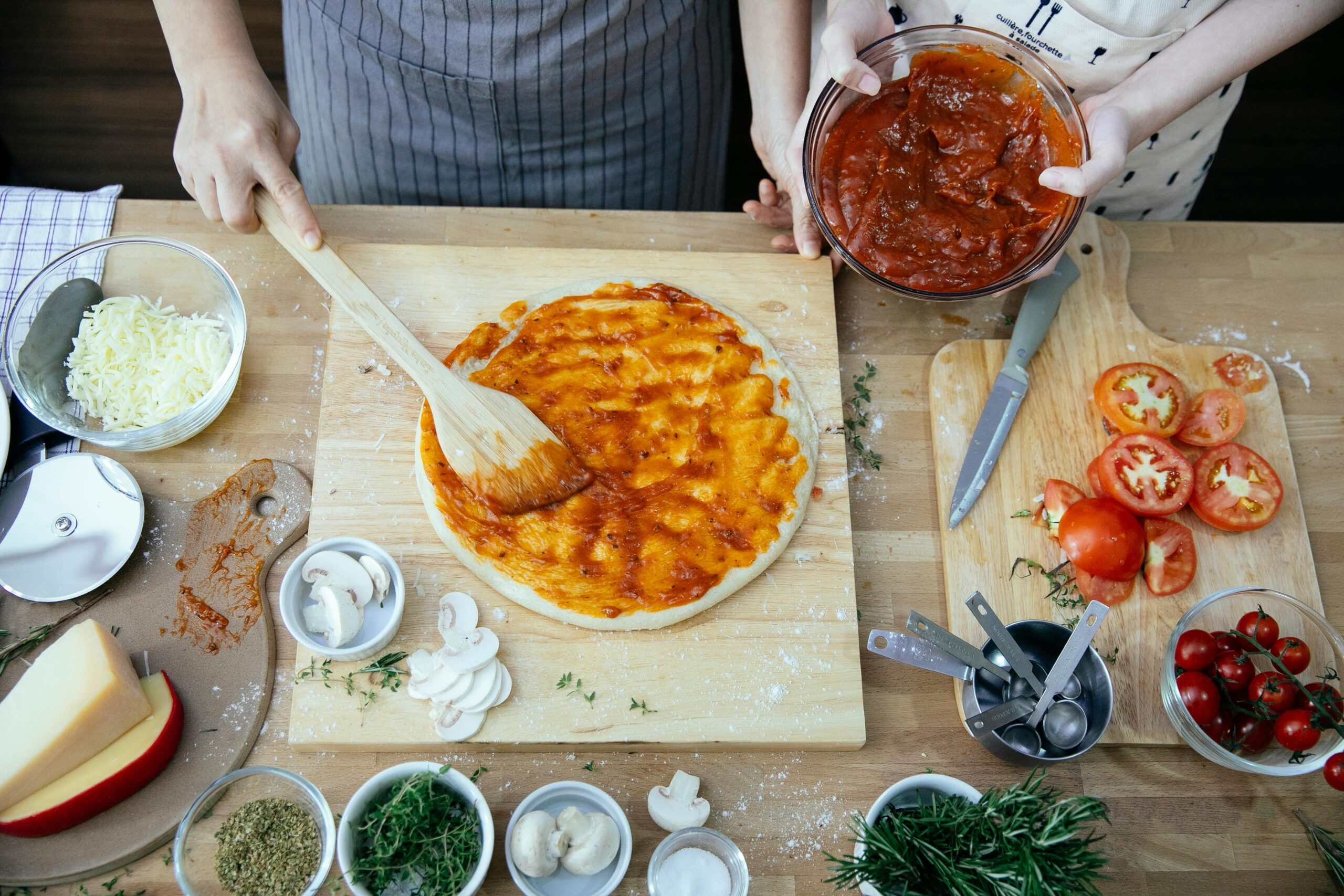 Hands preparing homemade pizza with tomato sauce and fresh ingredients on a wooden board.
