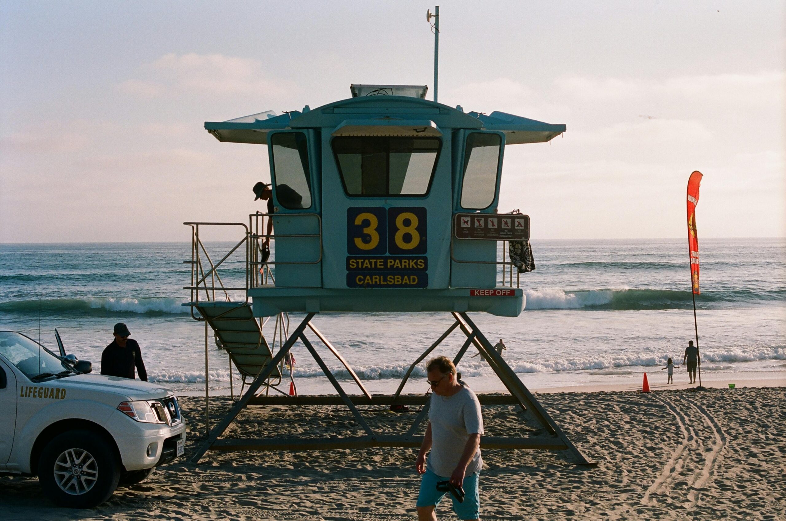 Lifeguard tower and vehicle on Carlsbad Beach with ocean waves and people enjoying a summer day.
